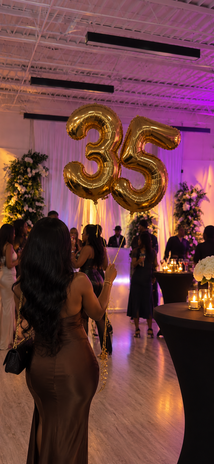 A guest holding gold number balloons at a Birthday Suite social night — purple uplighting, candlelit tables, and a full room celebrating